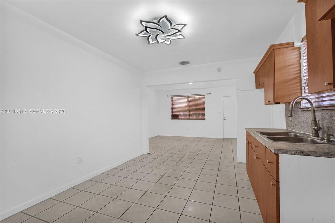 Kitchen featuring wood-toned cabinetry, a stone-finish countertop, dual-basin stainless steel sink with a high-arc faucet, neutral tile flooring, and a decorative multi-petaled flush-mount light fixture