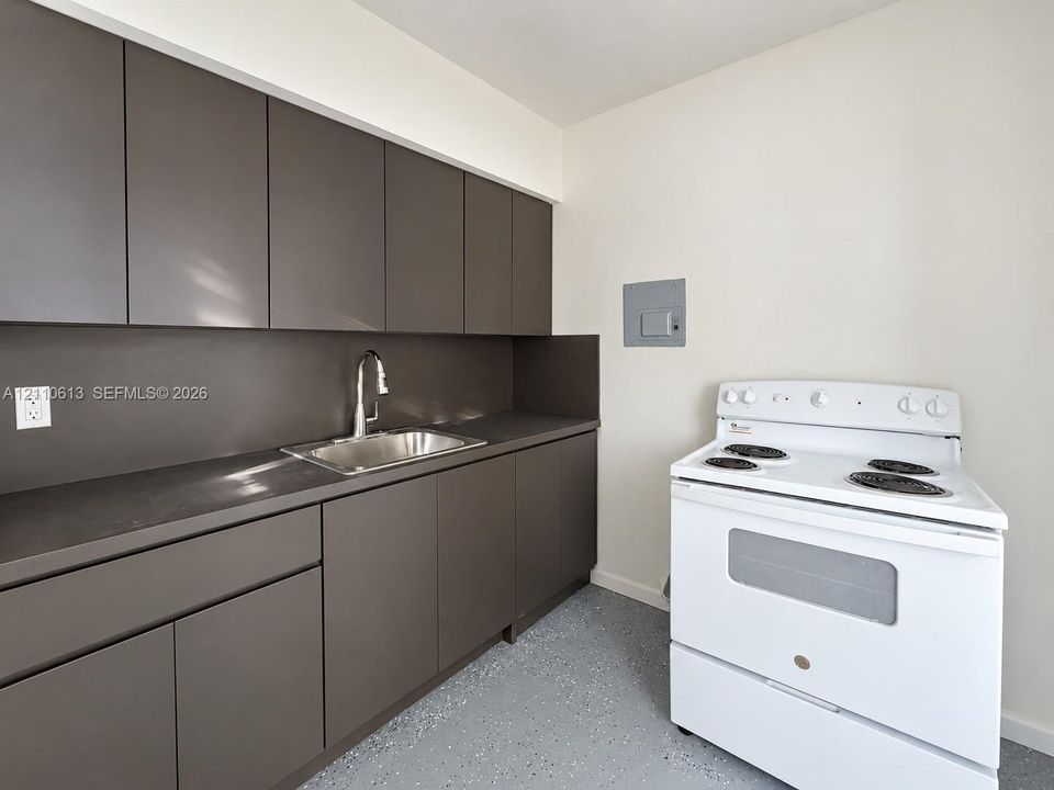 Dark-toned flat-panel cabinetry, matte dark countertops, stainless steel sink with pull-down faucet, white electric range, and speckled epoxy flooring