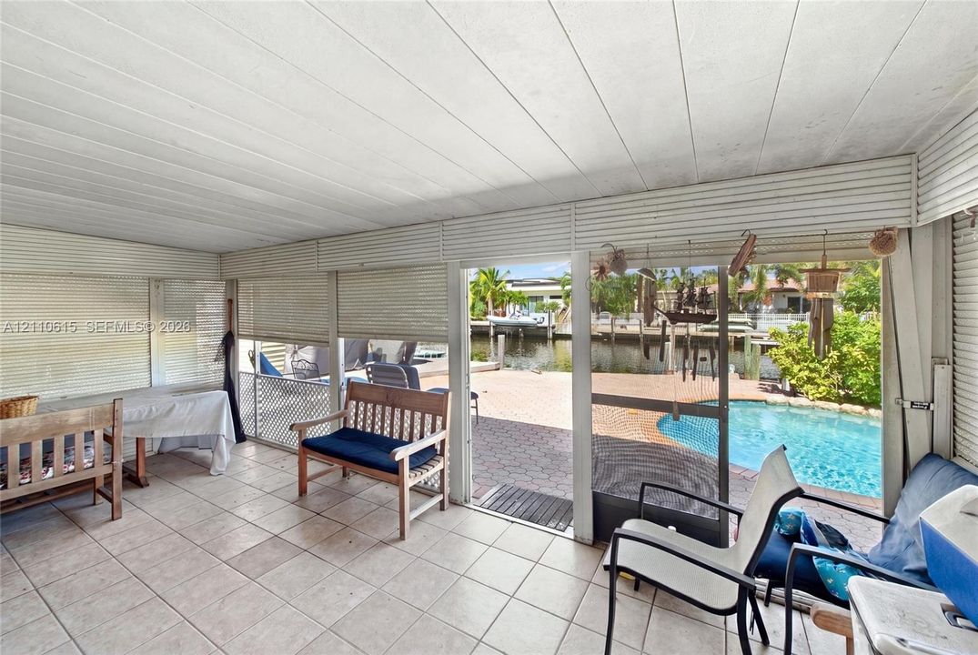 Enclosed patio featuring tile flooring, horizontal slat wall finishes, ceiling panels, and direct access to a paved pool deck with a waterfront bulkhead