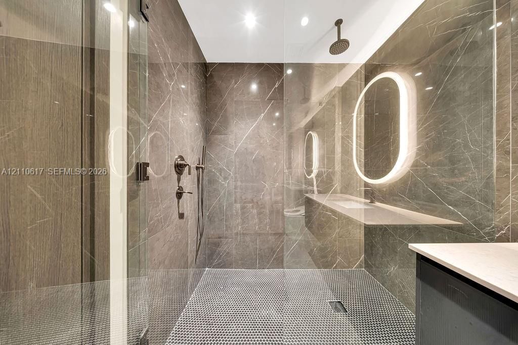 Walk-in shower featuring floor-to-ceiling stone-finish tile, a rainfall showerhead, a frameless glass enclosure, a penny-tile floor, and an integrated illuminated oval mirror