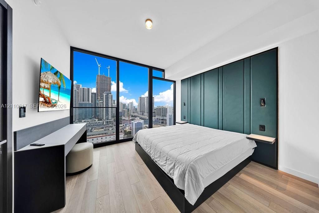 Floor-to-ceiling windows with urban skyline backdrop, custom floor-to-ceiling upholstered teal headboard, built-in desk with integrated shelving, and wide-plank wood-finish flooring