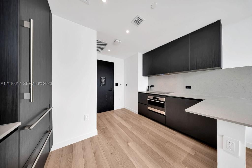 Kitchen featuring flat-panel dark cabinetry, integrated refrigerator, neutral solid-surface countertops, recessed lighting, and wood-finish flooring
