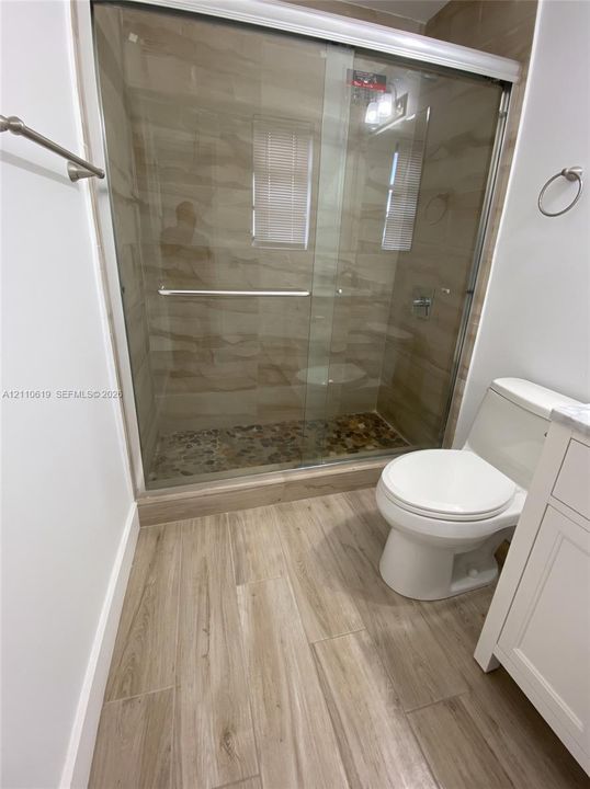 Walk-in shower with sliding glass doors, river stone flooring, and tiled walls, featuring wood-finish flooring and a white vanity