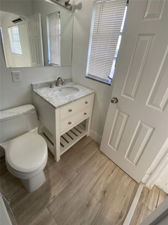 Bathroom featuring a vanity with stone-finish countertop, undermount sink, two-drawer storage, and slatted lower shelving, complemented by wood-finish flooring and a wall-mounted mirror