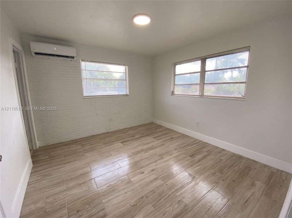 Interior room featuring painted brick wall detailing, wood-finish tile flooring, dual-pane windows with horizontal blinds, and a wall-mounted ductless climate control unit