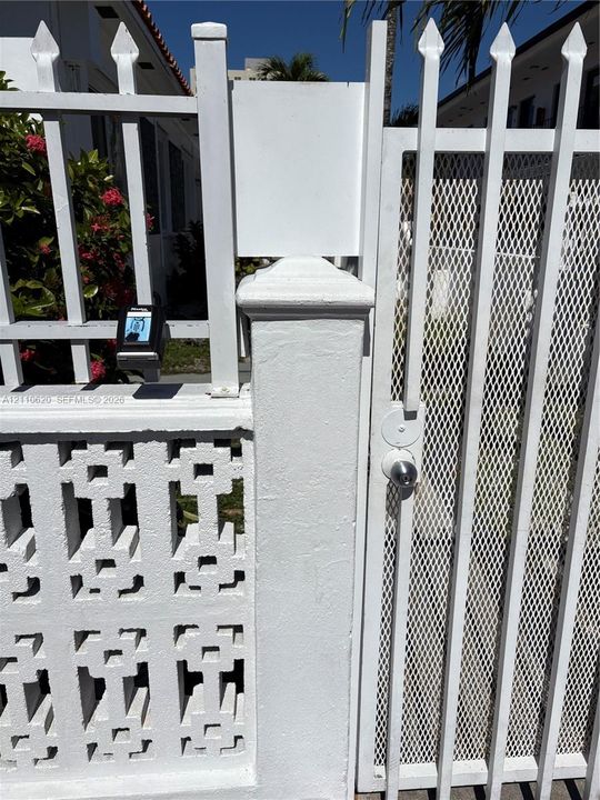 White metal pedestrian gate with spear-top finials, diamond-pattern wire mesh screen, and keyed entry, integrated into a white decorative concrete block wall