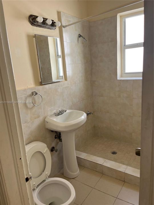 Bathroom featuring a pedestal sink, walk-in shower with tiled surround, tile flooring, wall-mounted light fixture, and framed mirror