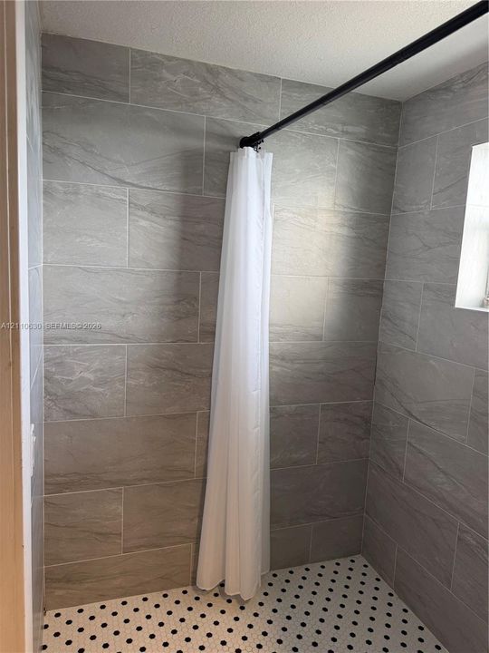 Walk-in shower enclosure featuring large-format grey tile wall surrounds, hexagonal floor tile with black accent detailing, and a black curtain rod