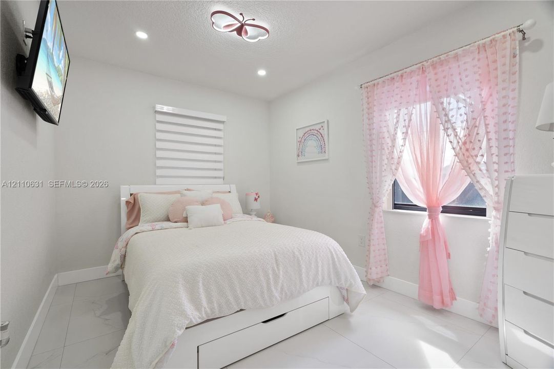 Bright bedroom featuring neutral tile flooring, recessed lighting, a stylized overhead fixture, and dual-shade window coverings
