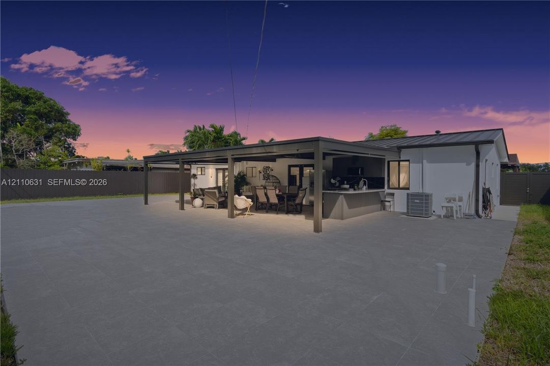 Expansive patio featuring large-format stone pavers, covered outdoor kitchen with integrated counter space, and dark-toned metal roof structure enclosed by privacy fencing
