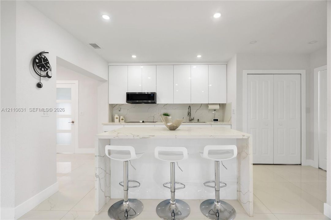 Kitchen featuring white high-gloss flat-panel cabinetry, waterfall-edge island with stone-finish countertop, recessed ceiling lighting, and tile flooring