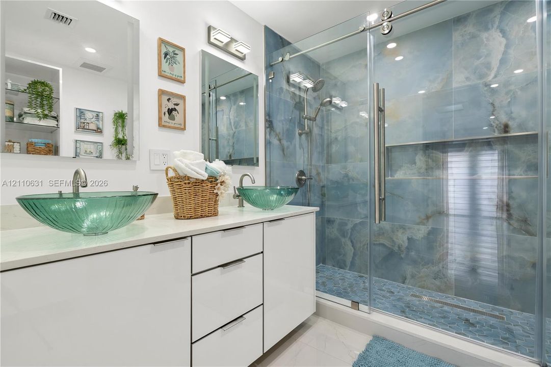 Double vanity featuring white cabinetry, quartz countertop, glass vessel sinks, and an expansive walk-in shower with floor-to-ceiling stone-patterned tile and a glass sliding door