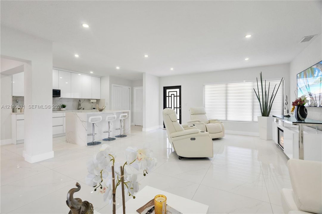 Open-concept living space featuring high-gloss cabinetry, stone-finish kitchen island with waterfall edge, recessed lighting, and large-format tile flooring