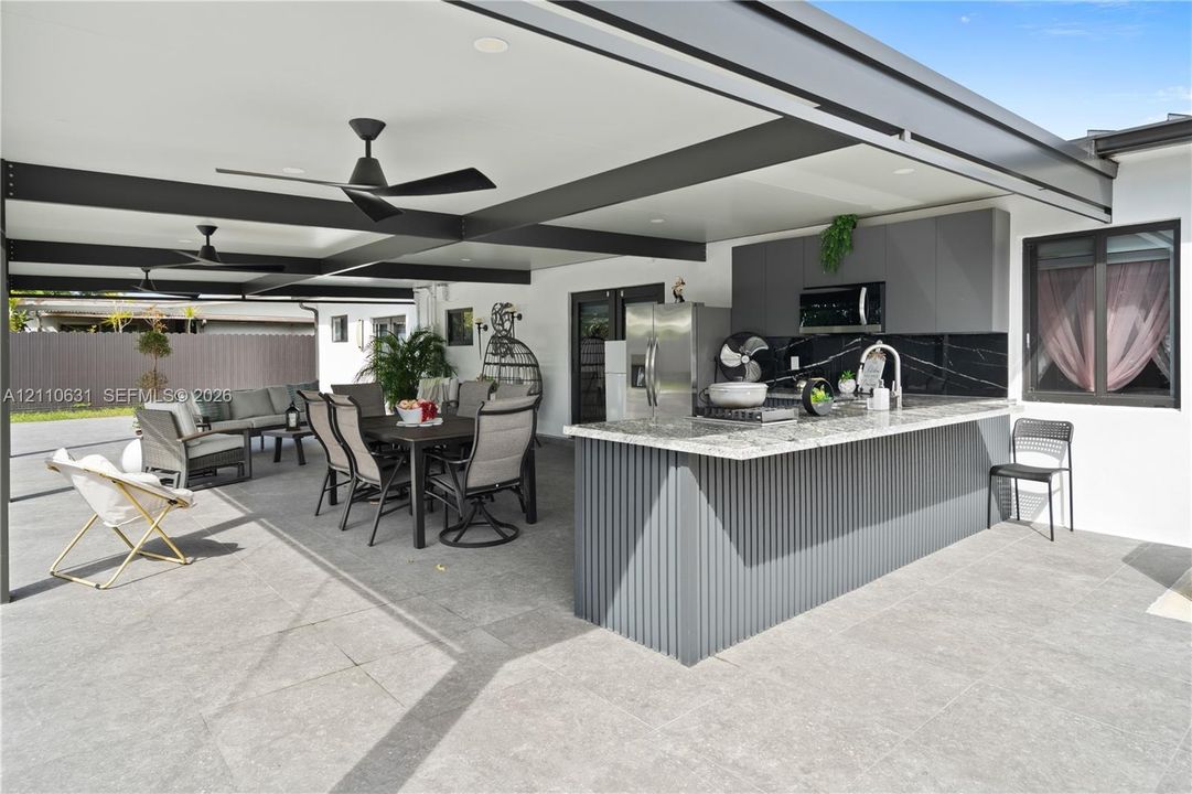 Covered outdoor kitchen featuring a stone-topped island with a fluted facade, dark cabinetry, high-contrast backsplash, and integrated ceiling fans
