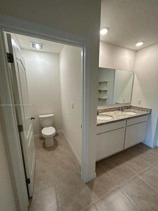 Dual-sink vanity with stone-finish countertop, white cabinetry, wall-to-wall mirror, and tile flooring