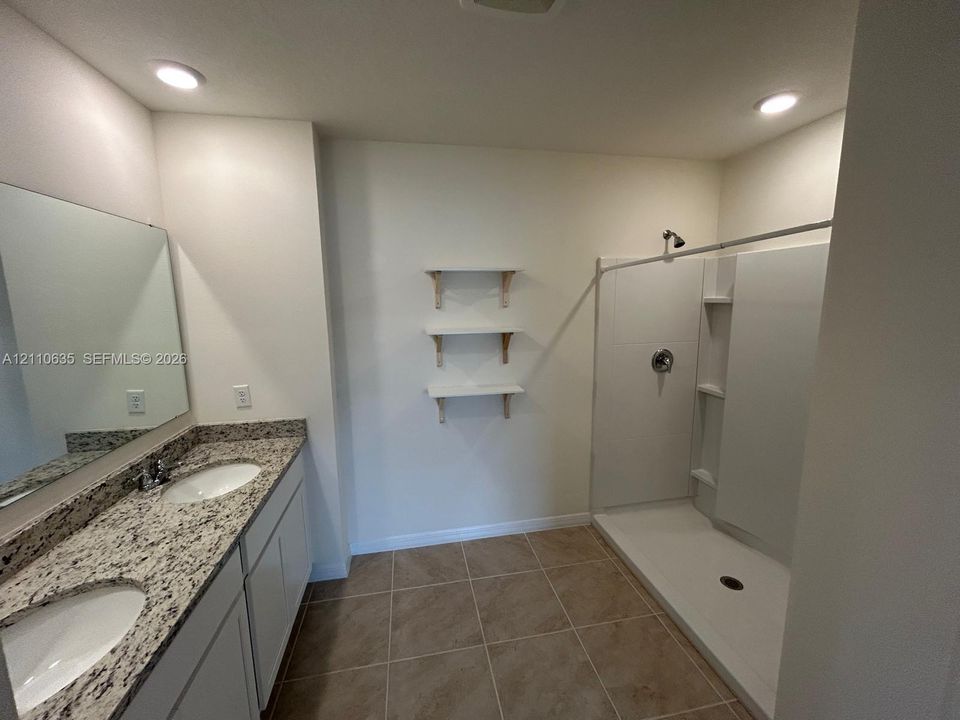 Bathroom featuring a dual-sink vanity with a stone-finish countertop, walk-in shower with integrated shelving, tile flooring, and wall-mounted wooden display shelves