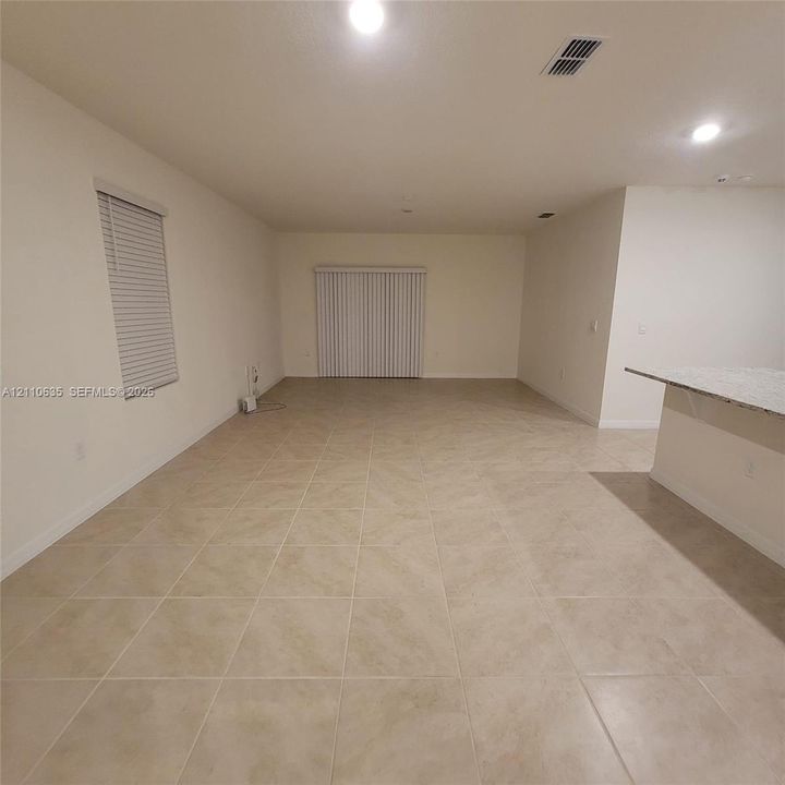 Open-concept space featuring large-format neutral tile flooring, recessed lighting, sliding glass doors with vertical blinds, and a kitchen peninsula with a light-toned stone-finish countertop