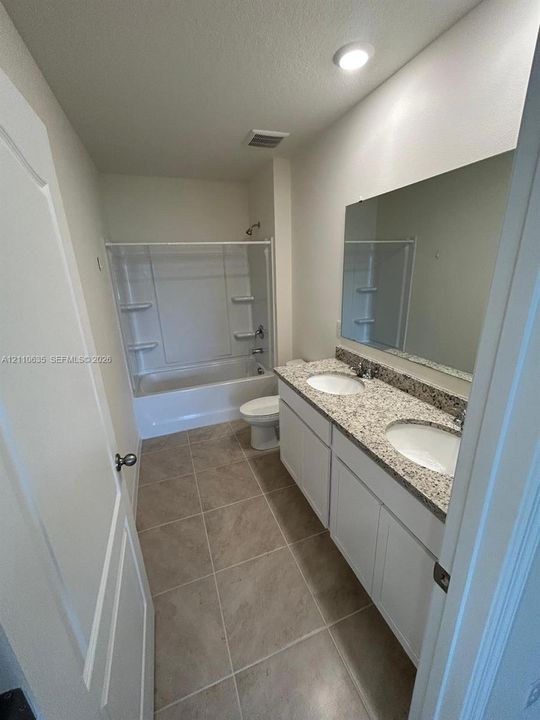 Dual-sink vanity with stone-finish countertop, white shaker-style cabinetry, large wall-mounted mirror, ceramic tile flooring, and tub-shower combination