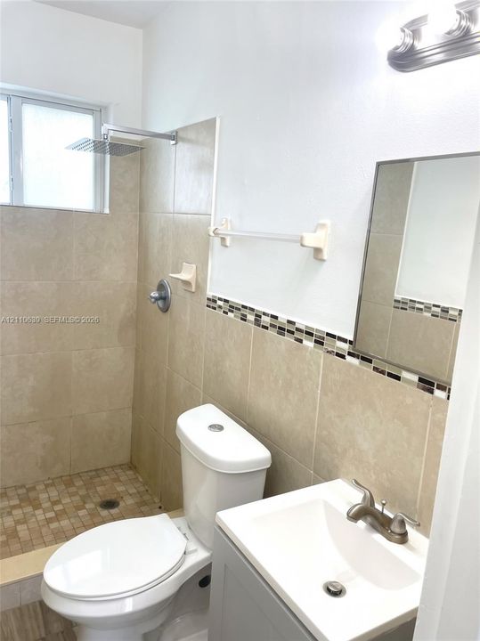 Bathroom featuring a shower with a rainfall showerhead, neutral tile surround with glass mosaic accent detailing, pedestal sink with metal fixtures, and a framed mirror