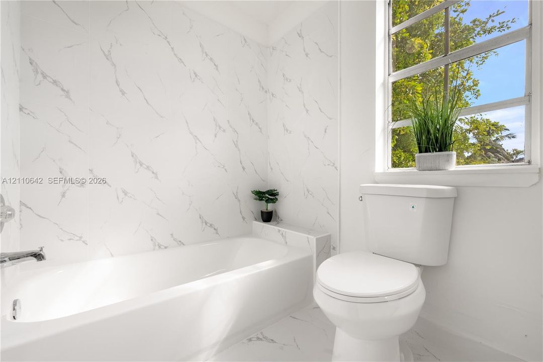 Full-height stone-patterned tile wall surround, integrated bathtub with polished chrome fixtures, white ceramic toilet, and large window with exterior greenery