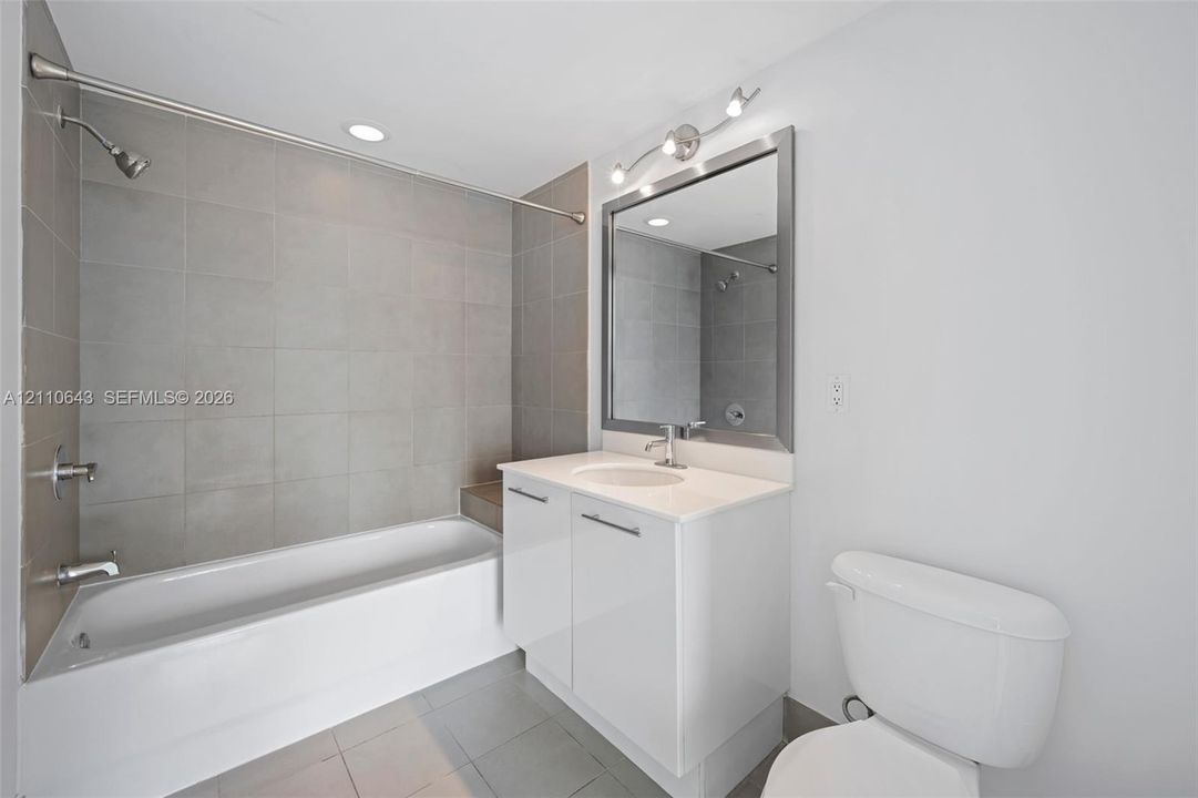 Bathroom featuring a porcelain tile bathtub surround, white vanity cabinet with an integrated sink, polished chrome faucet, framed mirror with integrated lighting, and neutral floor tile