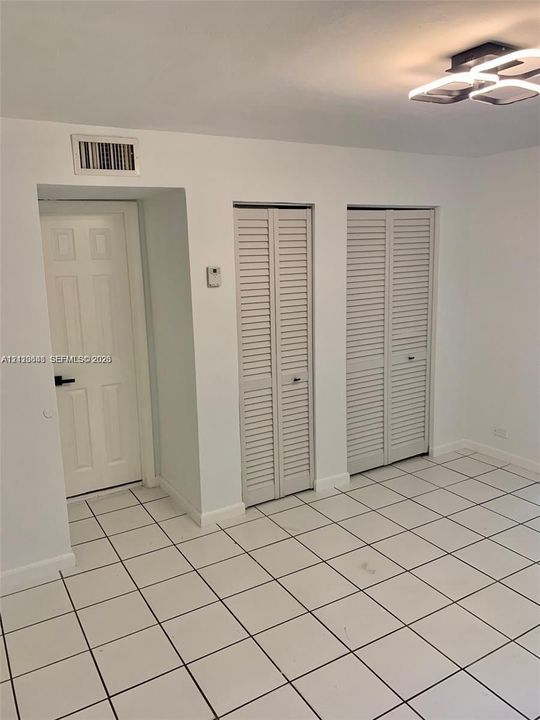 Neutral-toned room featuring large-format square tile flooring, two sets of louvered bifold closet doors, a single-panel interior door with dark hardware, and a modern geometric flush-mount ceiling light