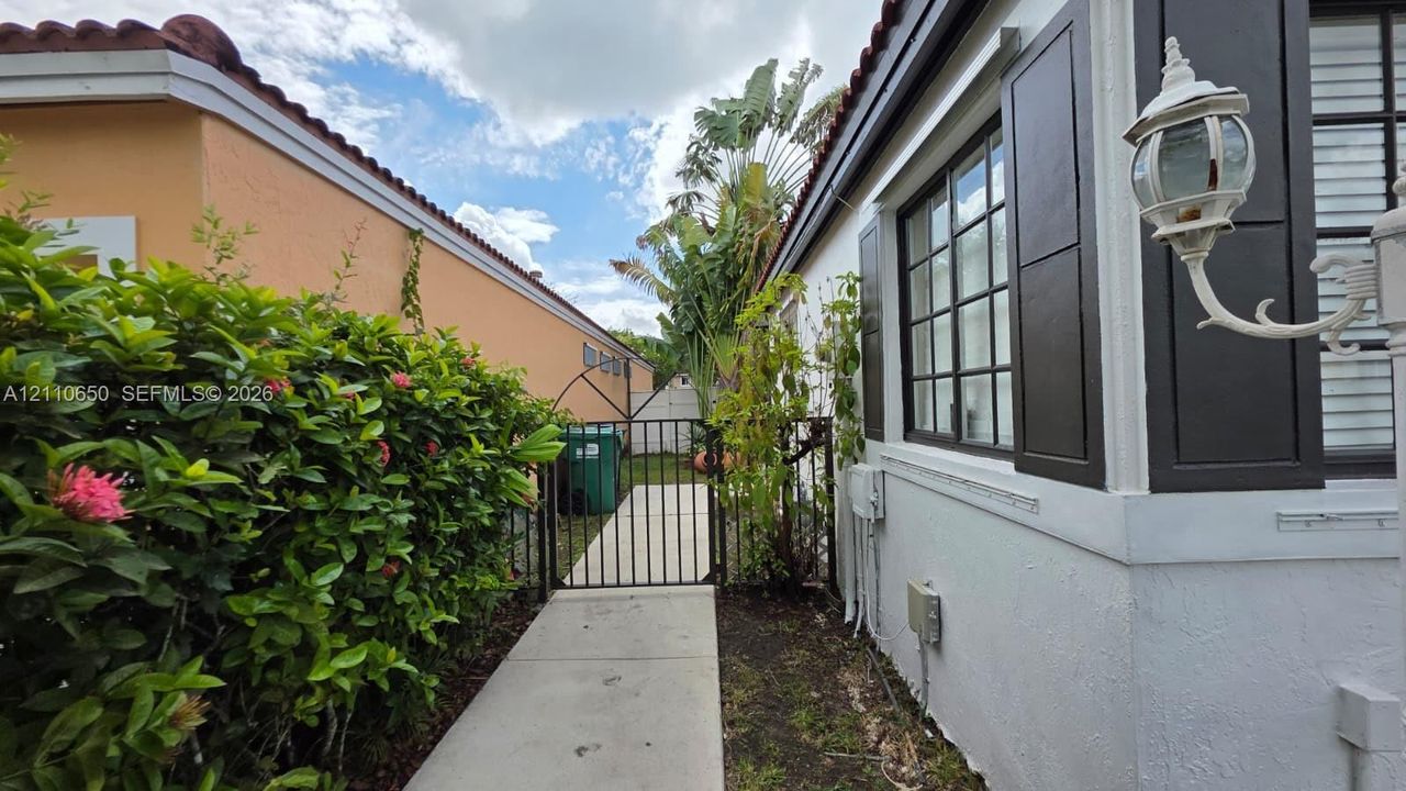 Concrete side walkway featuring decorative iron fencing, a gated entry, exterior window shutters, and established perimeter hedging