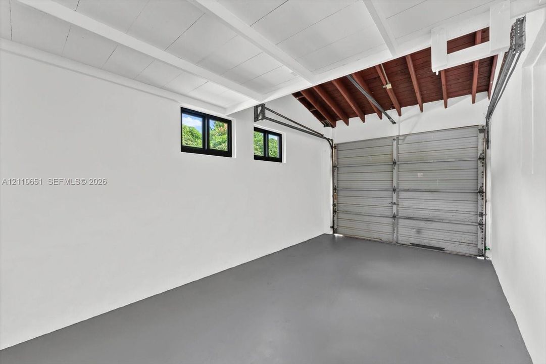 Garage space featuring painted concrete flooring, white walls, dual windows, and an exposed wooden beam ceiling
