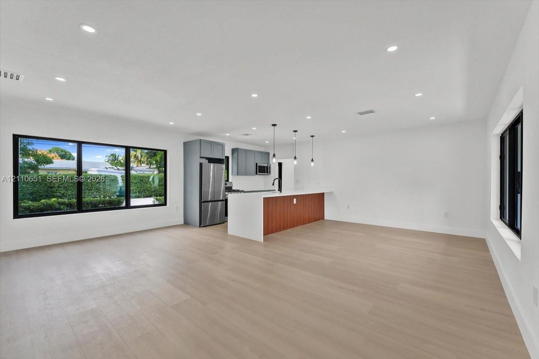 Open-concept interior featuring light wood-finish flooring, recessed lighting, kitchen island with stone-finish countertop, stainless steel refrigerator, and large-format black-framed windows