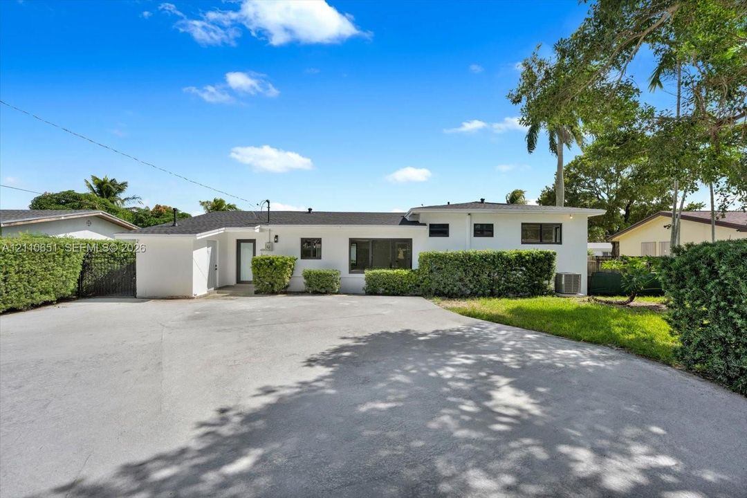 Single-story exterior featuring a painted stucco facade, asphalt shingle roof, expansive paved driveway, and manicured perimeter hedges