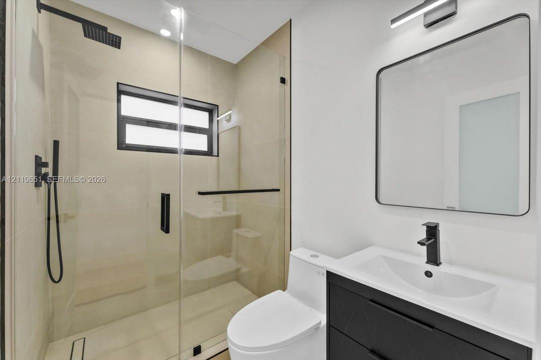 Bathroom featuring a glass-enclosed shower with a rainfall showerhead, a black vanity with a white countertop and rectangular sink, a black-framed mirror, and matte black fixtures