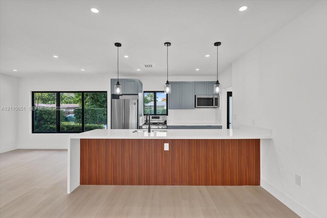 Kitchen featuring a waterfall-edge stone countertop, custom wood-slat cabinetry island, stainless steel appliances, matte black hardware, and wood-finish flooring