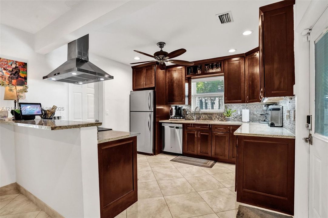 Kitchen featuring dark wood cabinetry, stone countertops, a mosaic tile backsplash, stainless steel appliances, and a ceiling-mounted stainless steel vent hood