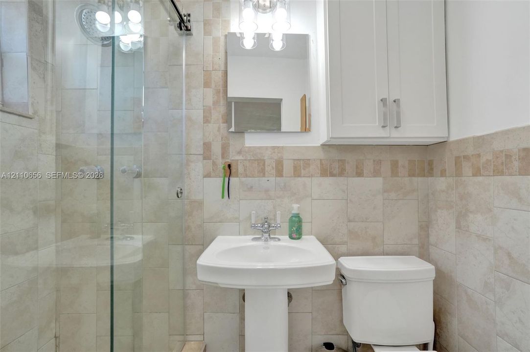 Glass-enclosed shower with stone tile surround, pedestal sink with polished chrome hardware, built-in white wall cabinetry, and coordinating stone wall tiling