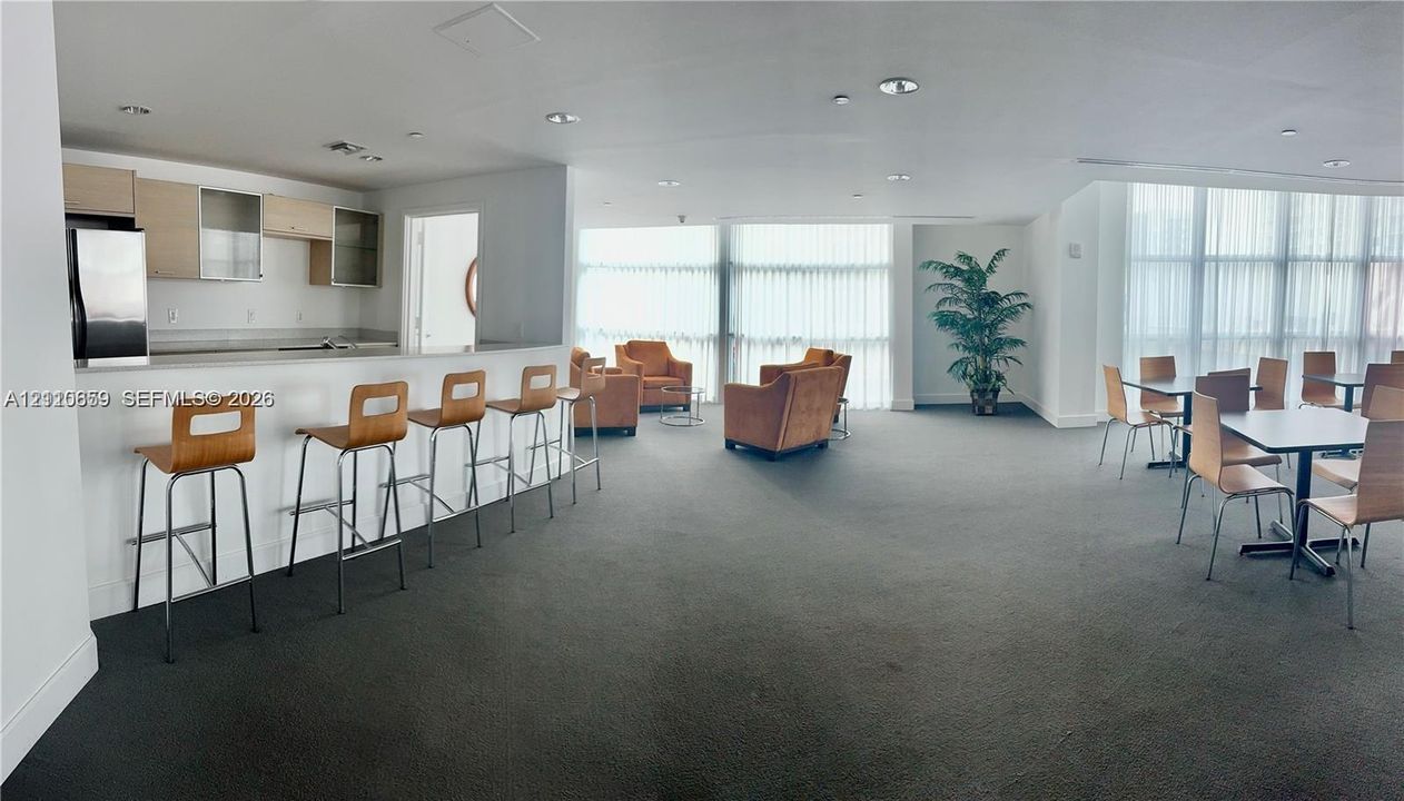 Open-concept common area featuring built-in kitchenette with light wood-finish cabinetry, bar-height counter seating, wall-to-wall carpeting, and floor-to-ceiling windows