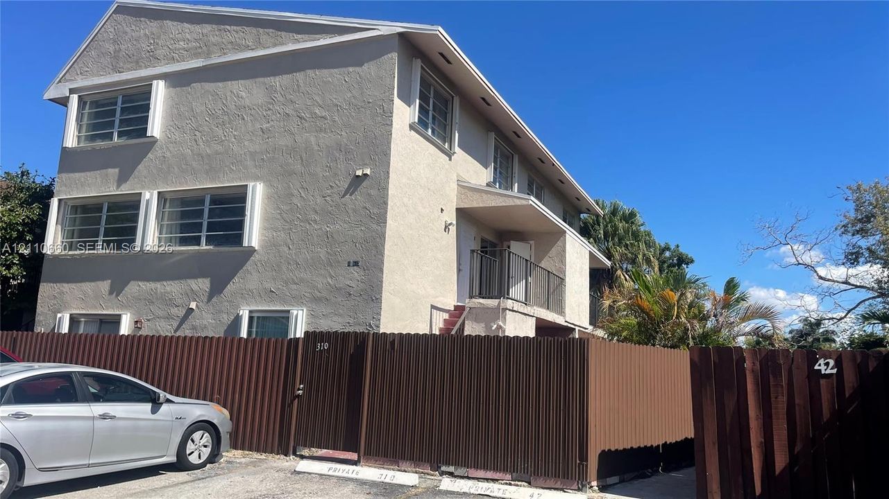 Multi-level building exterior with textured stucco siding, horizontal sliding windows, a private second-story balcony with metal railing, and a perimeter corrugated metal fence