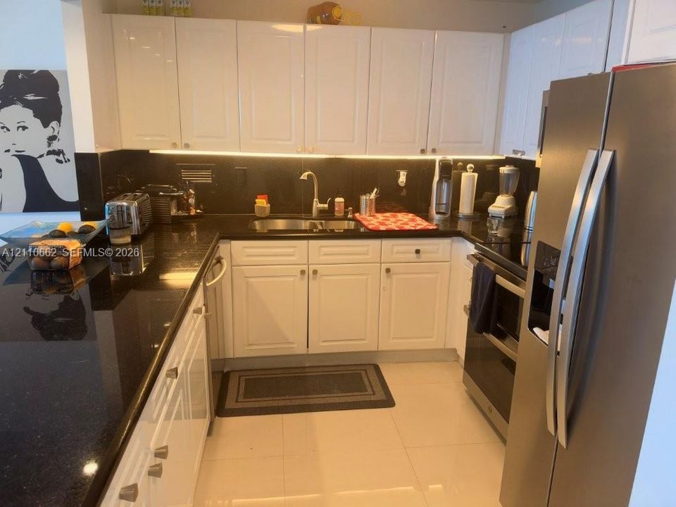 Kitchen featuring white shaker-style cabinetry, dark stone countertops, stainless steel appliances, under-cabinet lighting, and large-format tile flooring