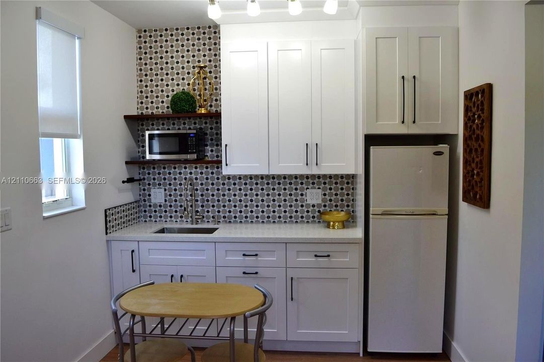 Kitchenette featuring white cabinetry with matte black hardware, stone-finish countertops, geometric-patterned tile backsplash, open wood shelving, and integrated stainless steel sink