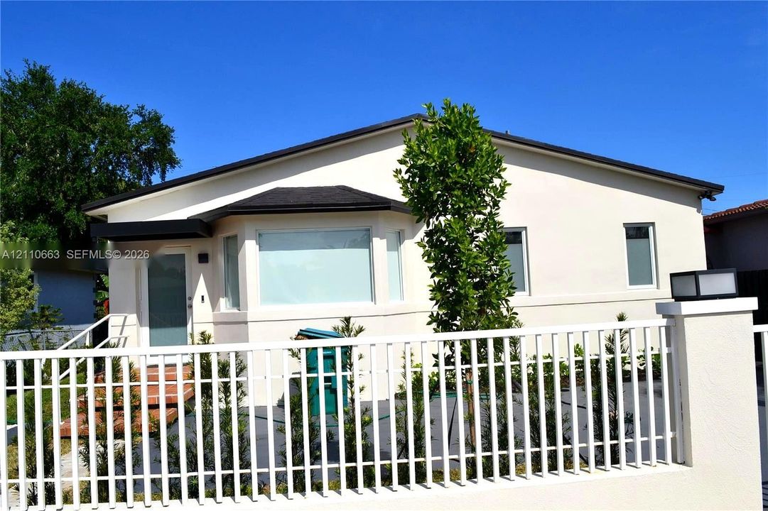 Stucco exterior with a bay window, shingle roofing, and a white picket-style fence