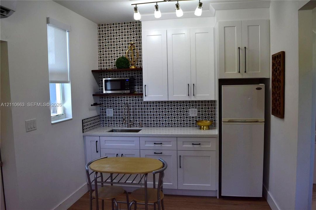 Kitchenette featuring white cabinetry with dark hardware, stone-finish countertops, geometric tile backsplash, open wood shelving, and integrated overhead track lighting
