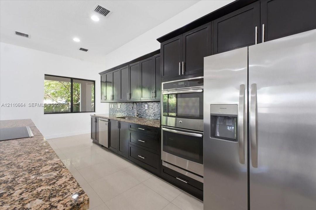 Spacious kitchen with a generous center island housing a new induction cooktop, granite countertops, rich dark cabinetry, double wall ovens, stainless steel refrigerator and dishwasher, recessed lighting, and tile flooring