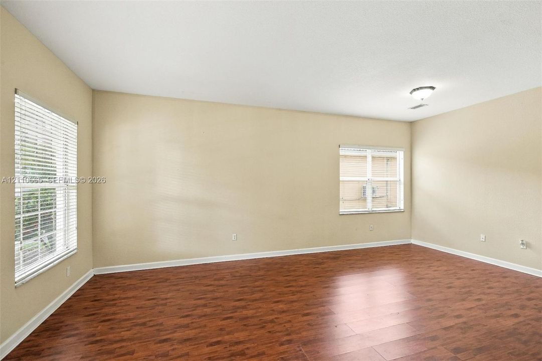 Spacious room featuring wood-finish flooring, neutral wall paint, two windows with horizontal blinds, and a flush-mount ceiling light fixture