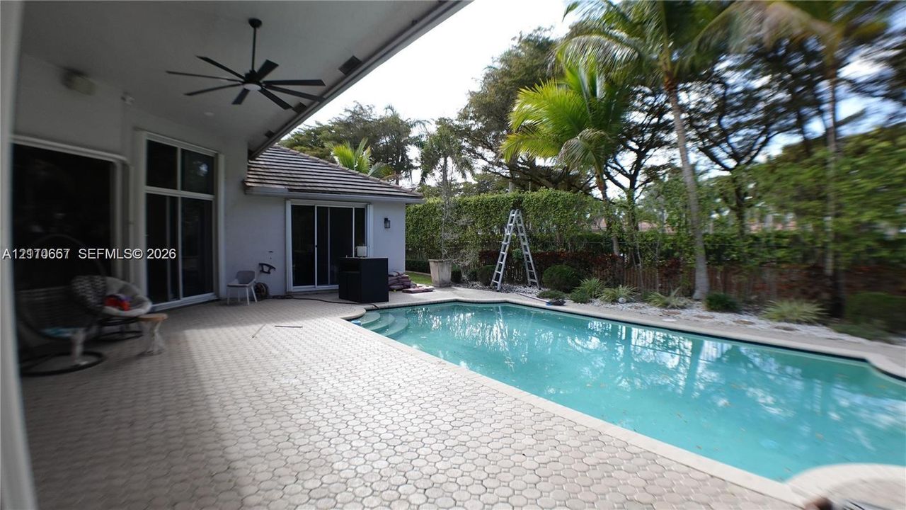 Covered patio featuring a patterned paver floor and ceiling-mounted fan, situated beside a rectangular swimming pool with integrated steps