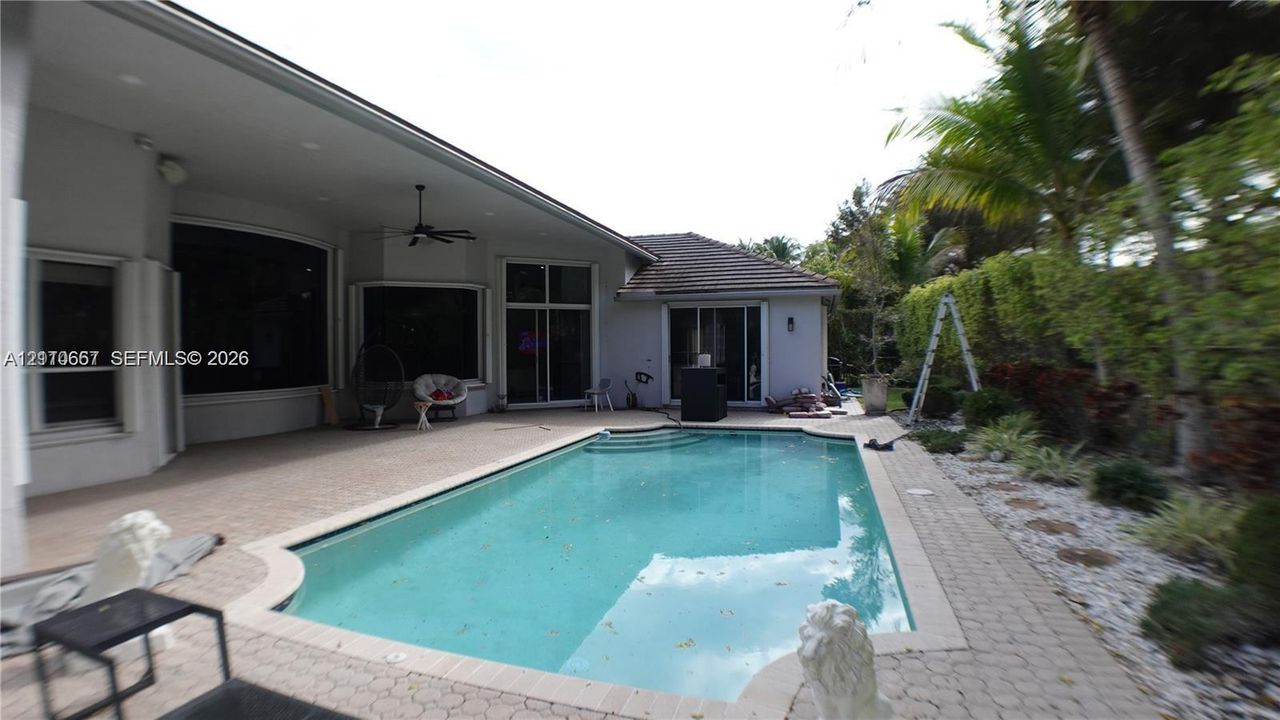 Covered patio with integrated ceiling fan, in-ground pool with stone paver decking, and mature tropical landscaping