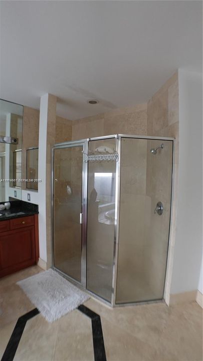 Corner shower enclosure with a metallic frame, neutral stone-finish wall tile, vanity with dark stone countertop, integrated mirror, and patterned tile flooring