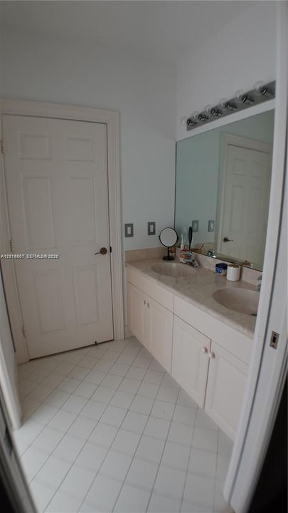 Double vanity featuring integrated sinks, stone-surface countertop, white cabinetry, large wall-mounted mirror, and tile flooring laid on a diagonal