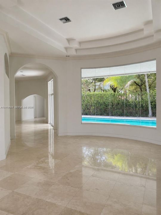 Interior featuring polished stone flooring, recessed tray ceiling, arched architectural detailing, and a expansive curved picture window showcasing a swimming pool