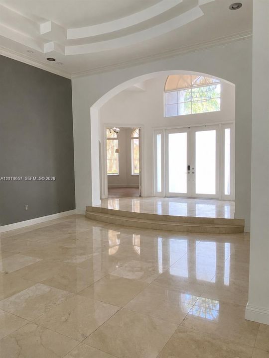 Grand foyer featuring polished stone flooring, a multi-tiered tray ceiling, an arched entryway, and double front doors with an overhead arched window
