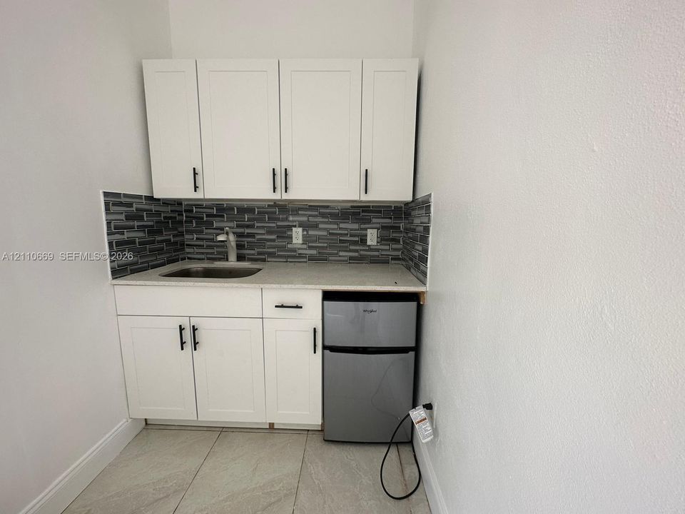 Kitchenette featuring white cabinetry with matte black hardware, stone-finish countertop, stainless steel sink with high-arc faucet, linear mosaic tile backsplash, and built-in under-counter refrigerator