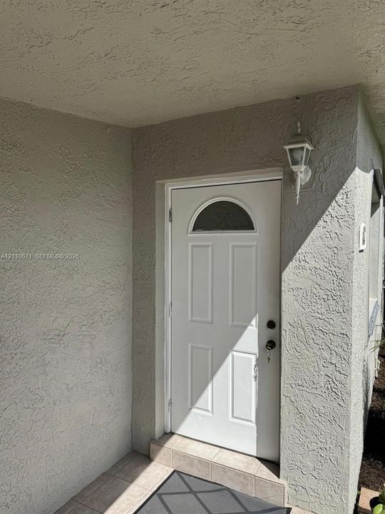 Covered entryway featuring textured stucco exterior, white six-panel door with arched glass insert, and mounted outdoor carriage light fixture